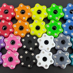 May include: A collection of colorful, star-shaped knobs in various colors, including red, orange, yellow, green, blue, purple, pink, black, white, gray, and teal. Each knob has a central hole and is adorned with small silver accents.