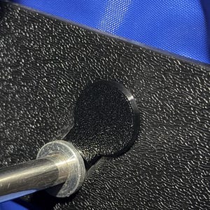 May include: Close-up of a black, textured surface with a circular, black knob attached to a silver metal rod. The surface has a repeating pattern. A blue fabric is visible in the background.