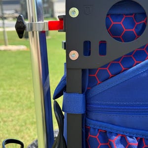 May include: Close-up of a black metal frame with a blue bag featuring a red honeycomb pattern. The frame has a circular cutout and is attached to a silver pole with a black knob. A red lever is visible. The bag has a blue strap.
