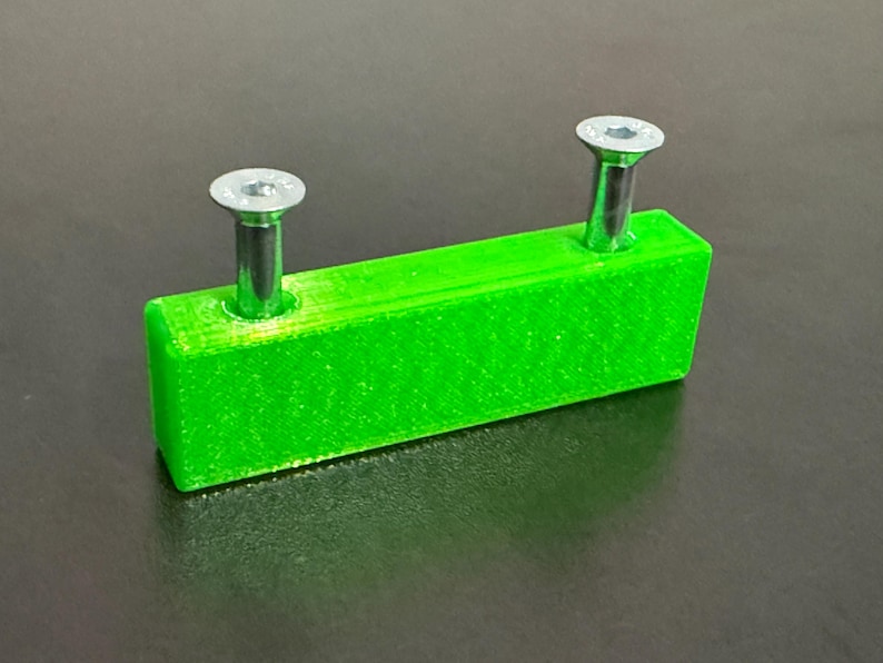 May include: A bright green rectangular object with two silver screws inserted. The screws have a flat, hexagonal head. The object is likely a component or part for a larger item.