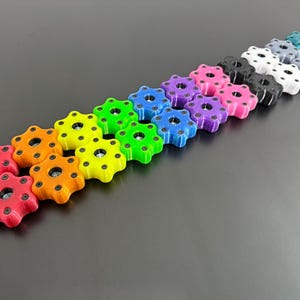 May include: A collection of colorful, star-shaped knobs in various hues, including red, orange, yellow, green, blue, purple, pink, black, white, gray, and teal. Each knob has a central hole and is arranged in a diagonal line against a dark gray background.