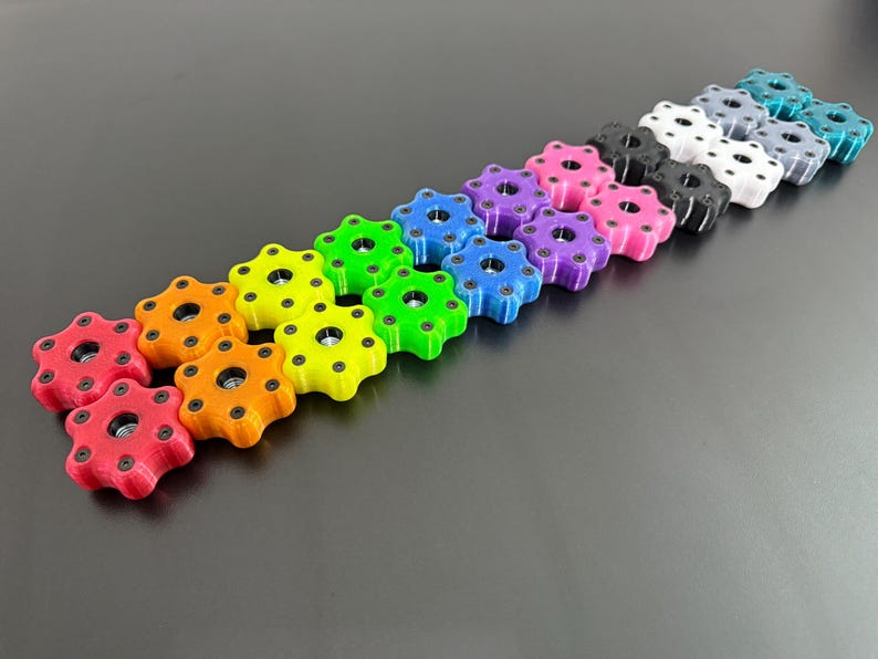 May include: A collection of colorful, star-shaped knobs in various hues, including red, orange, yellow, green, blue, purple, pink, black, white, gray, and teal. Each knob has a central hole and is arranged in a diagonal line against a dark gray background.