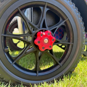 May include: Close-up of a black wheel with a red center cap. The wheel has a multi-spoke design and a black tire. The wheel is on a green grassy surface. The wheel is part of a mobility device.