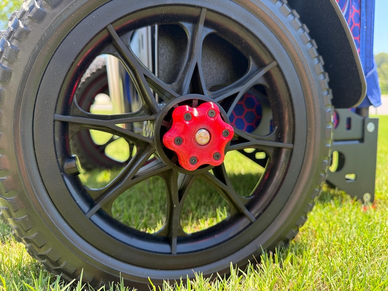 May include: Close-up of a black wheel with a red center cap. The wheel has a multi-spoke design and a black tire. The wheel is on a green grassy surface. The wheel is part of a mobility device.