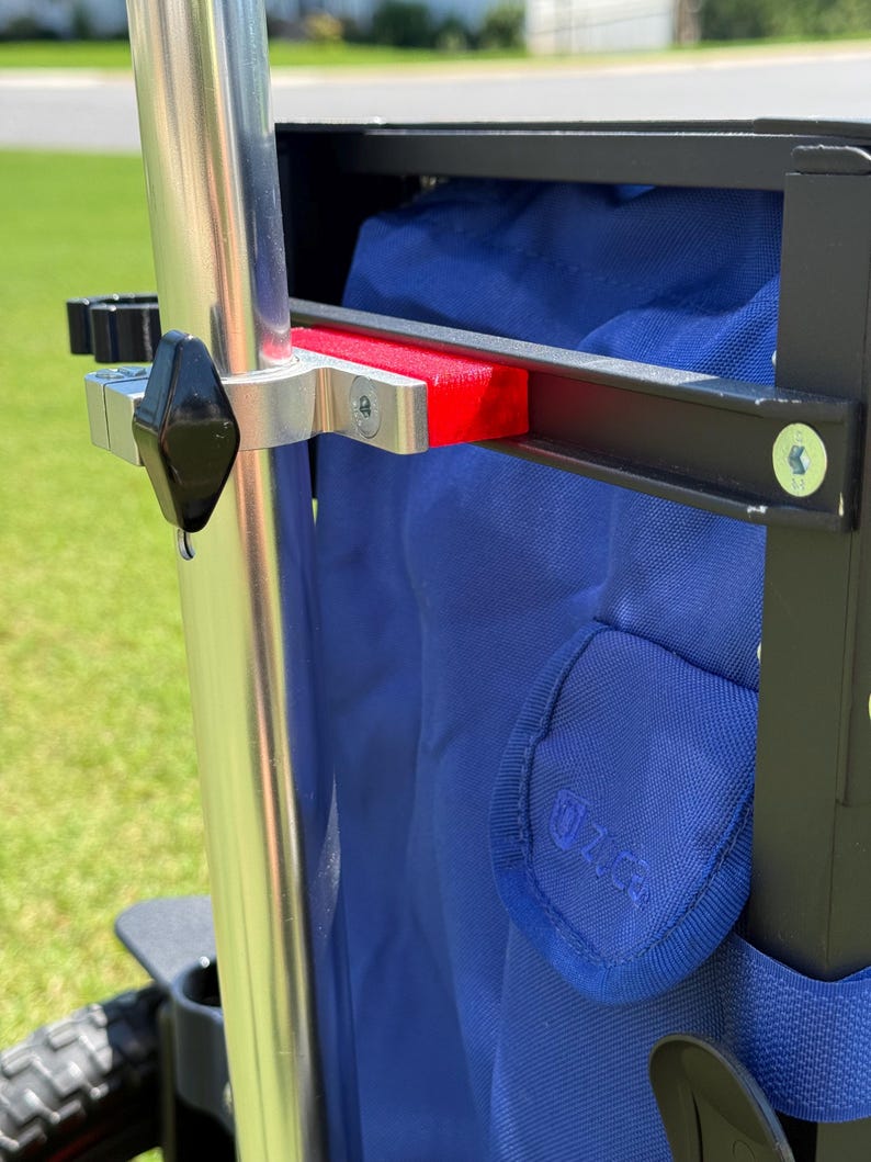 May include: Close-up of a utility wagon frame with a blue fabric interior. The frame is black with silver metal accents, including a vertical pole and a red support piece. The wagon has a black handle and black wheels.