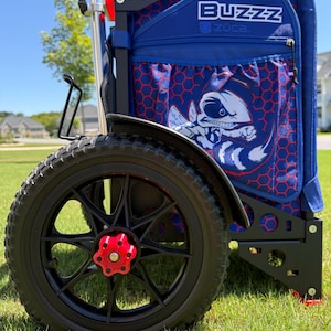 May include: A blue and red disc golf cart with the word "BUZZZ" on the side. The cart has a black frame, black wheels with red accents, and a graphic of a bee. The cart is on a grassy surface.