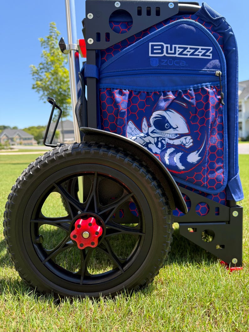 May include: A blue and red disc golf cart with the word "BUZZZ" on the side. The cart has a black frame, black wheels with red accents, and a graphic of a bee. The cart is on a grassy surface.