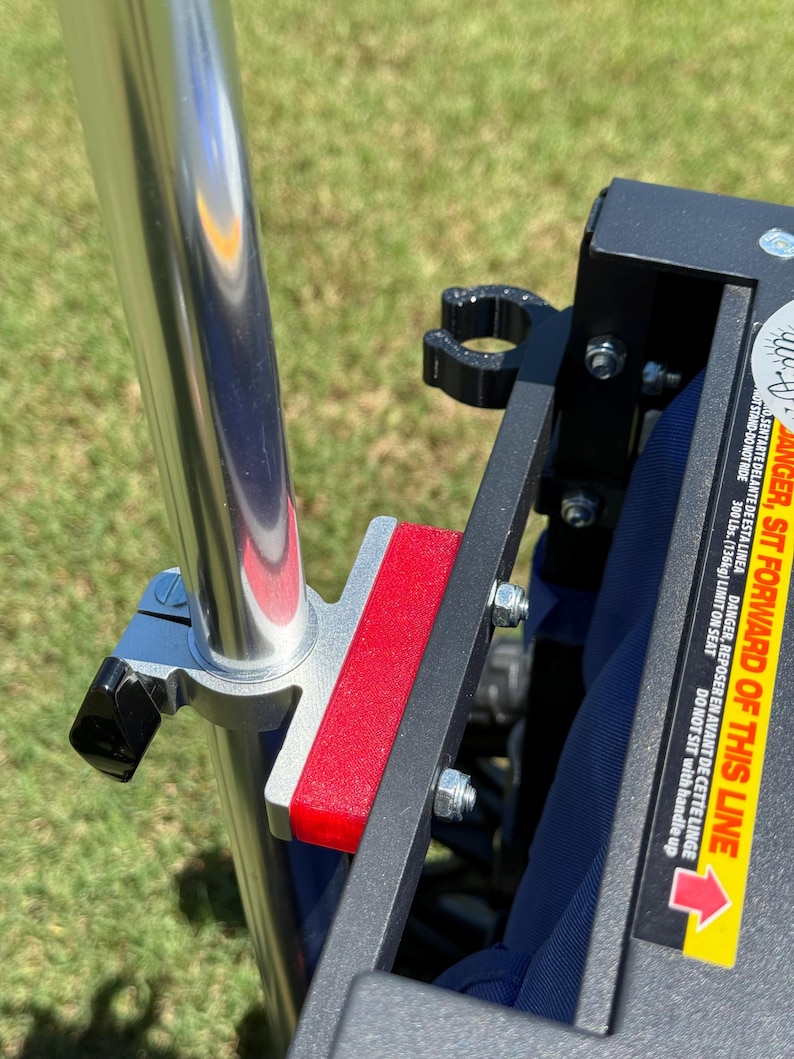 May include: Close-up of a metal frame with a red rectangular pad and a silver pole. The frame is attached with bolts and a black clamp. A warning label with text is visible.