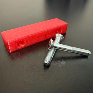 May include: A red rectangular object with two silver screws. The screws have a flat head and a threaded shaft. The red object has two holes on one side. The items are on a dark surface.