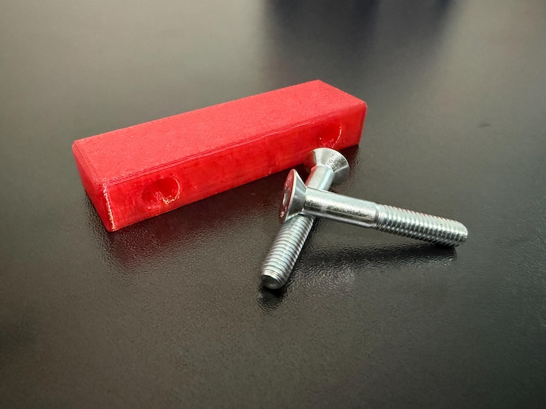 May include: A red rectangular object with two silver screws. The screws have a flat head and a threaded shaft. The red object has two holes on one side. The items are on a dark surface.