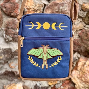 Könnte beinhalten: Blaue Umhängetasche mit braunem Riemen und Besatz. Die Tasche hat einen goldenen Reißverschluss und gestickte Designs, darunter eine grüne Motte, goldene Mondphasen und goldene Lorbeerblätter. Die Tasche ist aus Stoff.