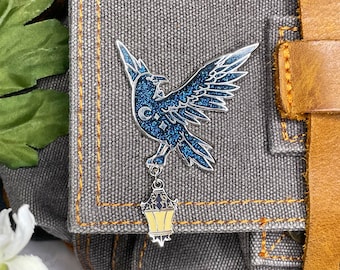 May include: A silver-toned raven pin with blue glitter wings and a crescent moon design. The raven is perched above a dangling lantern with a yellow and black design. The pin is set against a gray denim background.