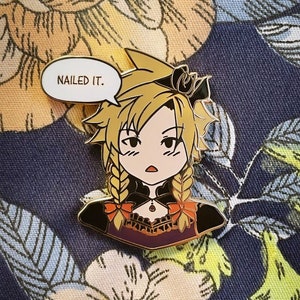 May include: A cartoon-style enamel pin featuring a character with blonde hair and braided pigtails. The character is wearing a black and purple outfit with orange accents. A speech bubble above the character reads "NAILED IT."