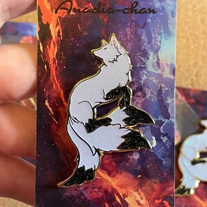 May include: A white and black enamel pin featuring a stylized fox with a glittery black tail. The pin is on a card with a watercolor background and the text "Anadia-chan" and "www.anadia-chan.com".