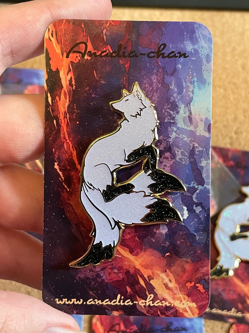 May include: A white and black enamel pin featuring a stylized fox with a glittery black tail. The pin is on a card with a watercolor background and the text "Anadia-chan" and "www.anadia-chan.com".