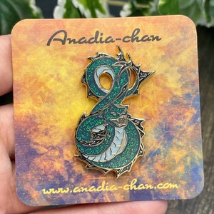 May include: A green and gold enamel pin featuring a dragon with glitter accents. The pin is on a card with the text "Anadia-chan" and "www.anadia-chan.com".