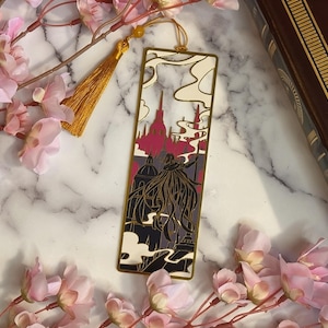 May include: A rectangular metal bookmark featuring a cutout design of a cloaked figure standing before a city skyline. The bookmark has a gold-toned frame, a color palette of cream, dark gray, and deep red, and includes a gold silk tassel at the top.