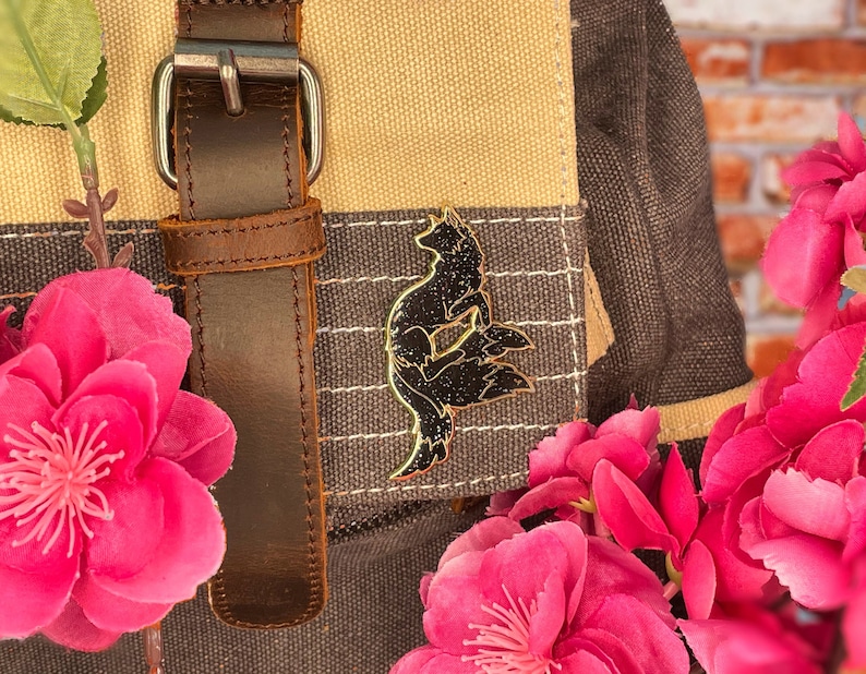 May include: A black metal pin shaped like a fox with a starry design on a brown canvas bag with a brown leather strap. The bag is decorated with pink flowers.
