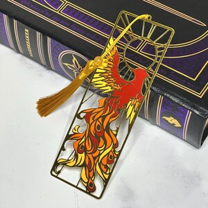 May include: A gold metal bookmark featuring a phoenix design in red, orange, and yellow. The bookmark has a tassel and is placed on a book with a black cover and gold accents. The book is titled "Warbreaker".
