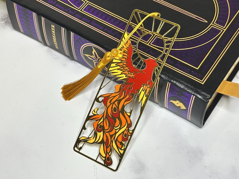 May include: A gold metal bookmark featuring a phoenix design in red, orange, and yellow. The bookmark has a tassel and is placed on a book with a black cover and gold accents. The book is titled "Warbreaker".