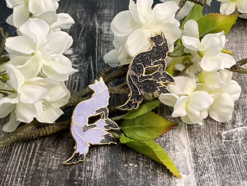 May include: Two enamel pins featuring a stylized fox design. The fox on the left is a light purple color with a black tail, while the fox on the right is black with a silver tail. Both foxes are sitting with their tails curled around their bodies.
