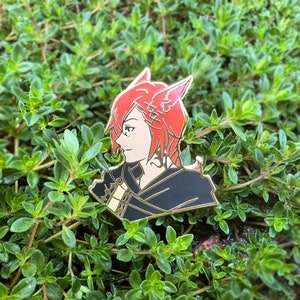 Könnte beinhalten: Detaillierter Emaille-Pin mit einer Figur mit roten Haaren und Katzenohren. Der Pin hat einen schwarzen und goldenen Umriss, wobei das Gesicht der Figur im Profil dargestellt ist. Der Pin ist vor einem Hintergrund aus grünem Laub angebracht.