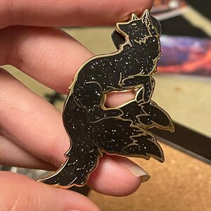 May include: A black enamel pin with a gold outline, depicting a stylized fox with a glittery finish. The fox is curled up with its tail wrapped around its body.