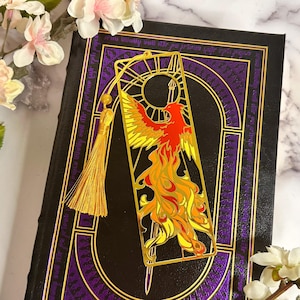 May include: A gold metal bookmark featuring a phoenix design in red, yellow, and orange, with a gold tassel. The bookmark is placed on a dark book with purple and gold accents.