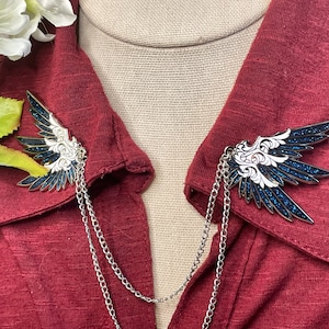 May include: Wing-shaped collar clips with a silver chain. The wings are detailed with white and dark blue enamel and glitter accents. The clips are displayed on a red garment.