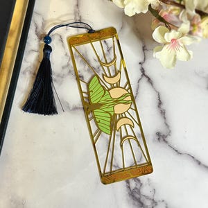 May include: A gold-colored metal bookmark featuring a green and cream-colored moth design. The bookmark has a rectangular shape with a decorative border and a navy blue tassel attached to the top. The bookmark is placed on a marble surface.