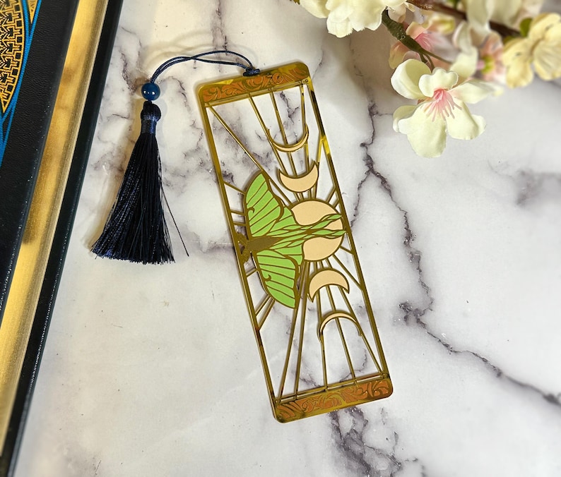 May include: A gold-colored metal bookmark featuring a green and cream-colored moth design. The bookmark has a rectangular shape with a decorative border and a navy blue tassel attached to the top. The bookmark is placed on a marble surface.