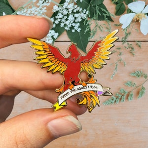 Puede incluir: Un pin de esmalte de fénix dorado con alas rojas, naranjas y amarillas. El fénix sostiene una pancarta que dice "From the ashes I rise".