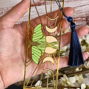 May include: Gold-colored metal bookmark featuring a green and cream-colored butterfly design, sun, and crescent moons. The bookmark has a navy blue tassel and a small blue bead. The bookmark is rectangular with a decorative border.