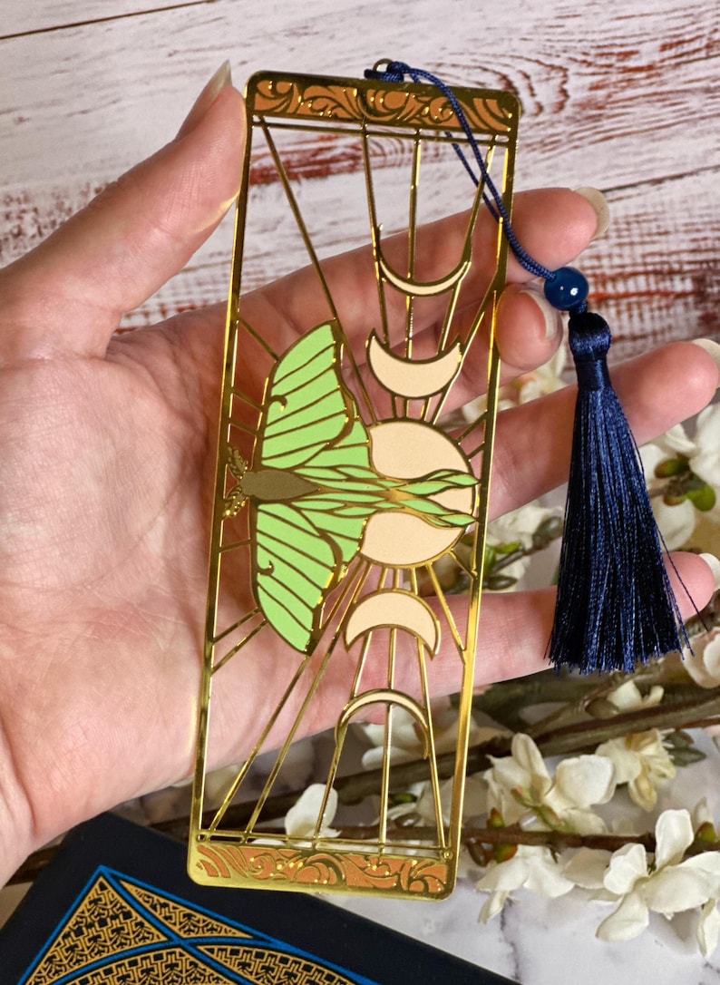 May include: Gold-colored metal bookmark featuring a green and cream-colored butterfly design, sun, and crescent moons. The bookmark has a navy blue tassel and a small blue bead. The bookmark is rectangular with a decorative border.