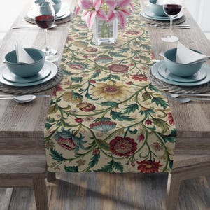 Puede incluir: Un camino de mesa floral con fondo crema y un estampado de flores rojas, amarillas y azules con hojas verdes. El camino de mesa está colocado sobre una mesa de madera puesta para una comida, con platos, vasos y cubiertos.