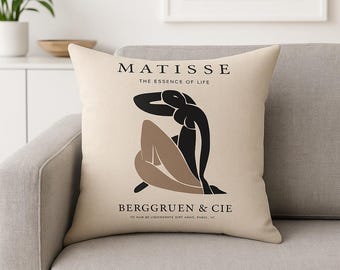 May include: Beige throw pillow featuring a Matisse-inspired design. The pillow displays a black and tan abstract figure with the text "MATISSE THE ESSENCE OF LIFE" and "BERGGRUEN & CIE" printed in black. The pillow is on a gray sofa.