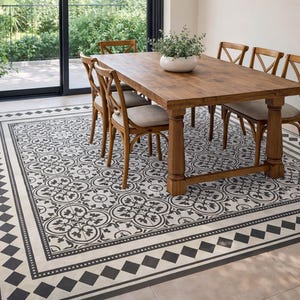 Op de afbeelding: Een eetkamer met een houten tafel en zes stoelen. De tafel staat op een patroonkleed met een zwart-wit bloemenontwerp. Een plant in een pot staat in de hoek en een glazen schuifdeur bevindt zich op de achtergrond.