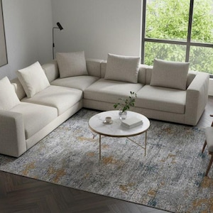 Puede incluir: Un salón moderno con un gran sofá seccional color crema y varios cojines. Una mesa de centro redonda con tapa de mármol descansa sobre una alfombra estampada. También hay un sillón y una planta en maceta.