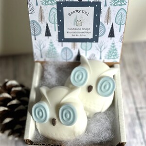 May include: Two white owl-shaped soaps with blue swirl eyes in a white box with a blue and white patterned lid. The lid has a label that says "Snowy Owl Handmade Soaps MountainhouseSoaps Net Wt. 4.0 oz."