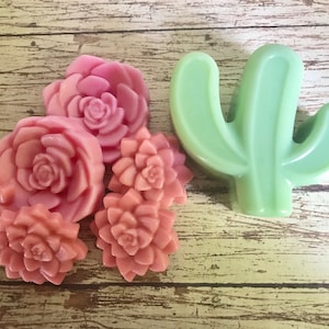 May include: Five pink and peach colored flower shaped soaps and a green cactus shaped soap. The soaps are arranged on a wooden surface.
