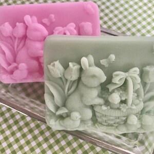 May include: Two handmade soap bars featuring a bunny rabbit design. One bar is pink with a bunny holding a basket of eggs. The other bar is green with a bunny holding a basket of flowers.