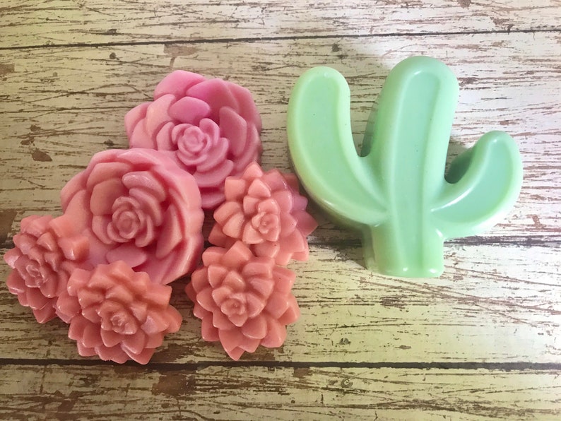 May include: Five pink and peach colored flower shaped soaps and a green cactus shaped soap. The soaps are arranged on a wooden surface.