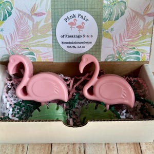 May include: A white gift box with pink flamingo shaped soaps inside. The box has a label that says "Pink Pair of Flamingo Soaps" and "MountainhouseSoaps" with a net weight of 5.4 oz.
