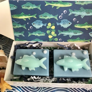 May include: Two blue and green fish-shaped soaps in a white box with a blue and green fish pattern on the inside of the lid. The box is filled with shredded paper.