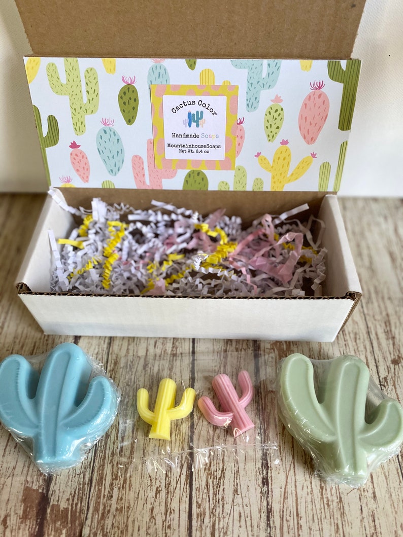 Pu&ograve; includere: Una scatola piena di carta triturata e tre saponi a forma di cactus. I saponi sono blu, verdi e gialli. La scatola &egrave; decorata con un motivo di cactus e un'etichetta che recita "Cactus Color Handmade Soaps MountainhouseSoaps Net Wt. 3.4 oz."