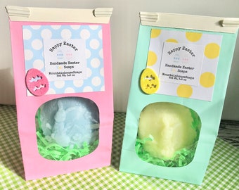 May include: Two pastel-colored gift bags with clear windows, each containing a handmade Easter soap shaped like an egg with a bunny design. The bags are decorated with polka dots and the text "Happy Easter" and "Handmade Easter Soaps".