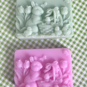 May include: Two rectangular soaps with an Easter theme. One soap is light green and the other is light purple. Both soaps feature a rabbit holding a basket of eggs and tulips.