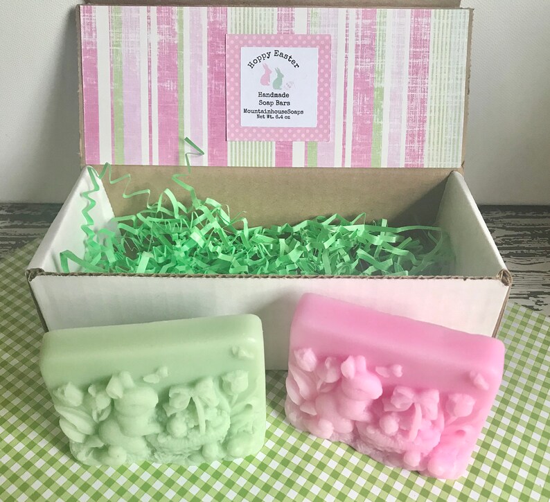 May include: Two handmade soap bars in a gift box. The soap bars are green and pink and feature a bunny rabbit design with the text "Hoppy Easter". The soap bars are packaged in a white box with green shredded paper. The box has a pink and green striped lid with a label that reads "Handmade Soap Bars MountainhouseSoaps Net Wt. 4 oz."