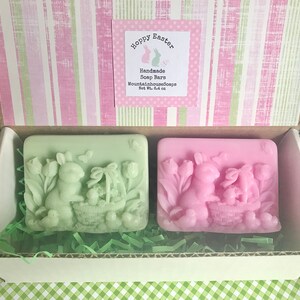 May include: Two handmade soap bars in a gift box. The soap bars are shaped like bunnies holding baskets of eggs. One bar is green and the other is pink. The box is decorated with pink and green stripes and a label that says "Hoppy Easter Handmade Soap Bars MountainhouseSoaps Net Wt. 6.4 oz."