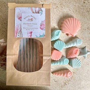 May include: A brown paper bag with a window showing a blue and white striped pattern. The bag has a label that says "Beach Walks Handmade Shell Soaps MountainhouseSoaps Net Wt. 3.5 oz".  The bag is surrounded by 10 handmade soap shells in shades of pink and blue, shaped like seashells and a starfish.